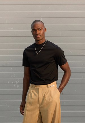 Μαύρο t-shirt με στρογγυλή λαιμόκοψη και κοντά μανίκια, συνδυασμένο με μπεζ πλισέ παντελόνια που διαθέτουν ετικέτα στη μέση και δύο κουμπιά στο πλάι.