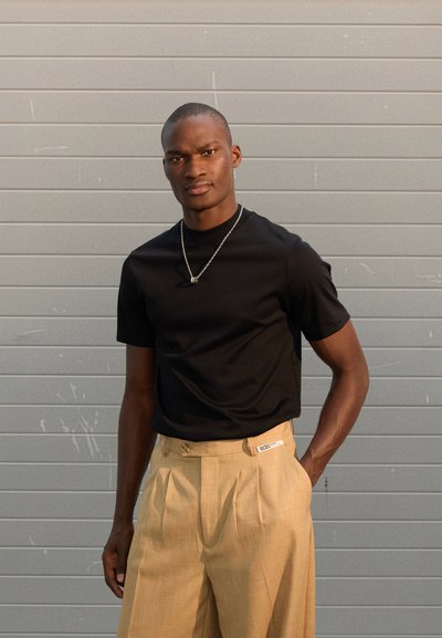Black crew neck t-shirt with short sleeves paired with beige pleated trousers featuring a label on the waistband and two side buttons.