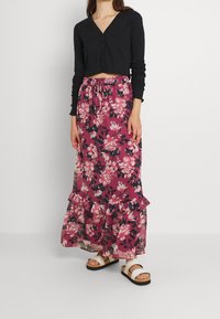 Cárdigan corto de canalé negro, falda maxi floral en tonos rosa y oscuros, con dobladillo acampanado, combinada con sandalias blancas de tiras.