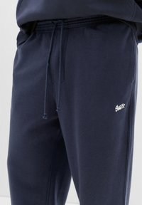 Námorníckom modré tepláky vyrobené z mäkkej tkaniny, so elastickým pásom na šnúrku, bočnými vreckami a malým bielym logom na stehne.