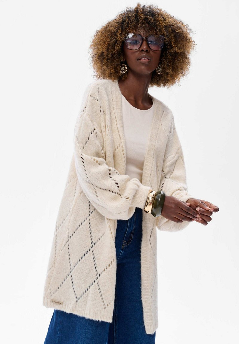 Beige gebreide cardigan met een diamanten patroon, lange mouwen en een open voorkant. Gestyled met een lichte top en wijde jeans.
