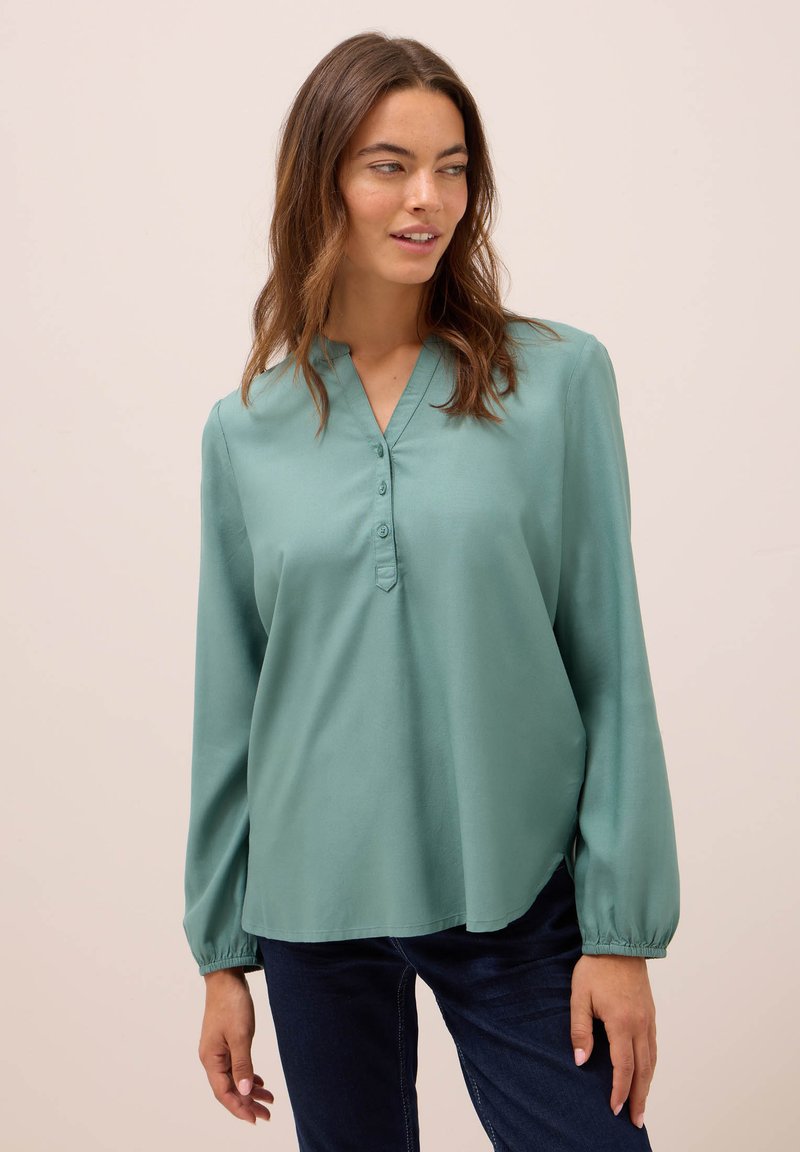 Kvinna i en lös, långärmad, tealblå blus med knappar vid kragen, ihop med mörkblå jeans, stående mot en neutral bakgrund.