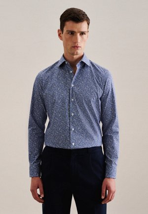 Camicia di cotone a maniche lunghe con motivo floreale blu, colletto a punta e bottoni bianchi. Indossata con pantaloni scuri. Tessuto morbido.