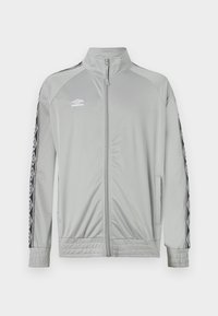 Chaqueta deportiva de cremallera en gris claro con cuello alto, rayas en blanco y negro en las mangas y el logotipo de Umbro en el lado izquierdo del pecho.