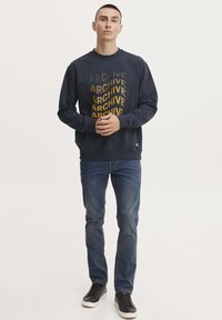 Sudadera navy con la palabra "ARCHIVE" impresa en filas verticales en amarillo y oliva. Combinada con jeans azul y zapatillas negras.