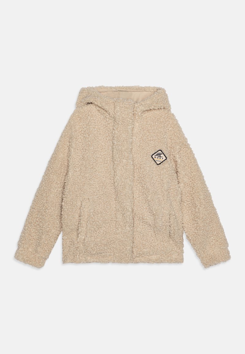 Veste en fausse fourrure beige avec une capuche, deux poches avant et un petit patch de logo noir et blanc sur le côté gauche. Texture douce.