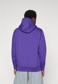 Purple hooded sweatshirt with a relaxed fit, ribbed cuffs, and hem. Smooth texture with minimal detailing, worn over light gray shorts.
