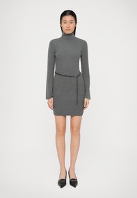Grå ribbet turtleneck kjole med en tætsiddende silhuet, der har lange ærmer og et sort kædebælte i taljen. Sorte spidse sko.