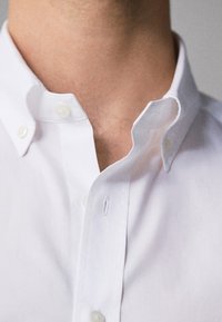 Close-up of a person wearing a white button-down shirt with the top button undone, showing part of their neck and collar.