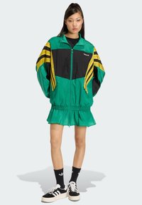 Zelená a čierna bunda na zips so žltými pruhmi, elastickým pásom a volánovým lemom. Nosená cez čierne tričko s čierno-bielymi teniskami.
