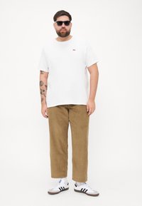 T-shirt en coton blanc avec un petit logo rouge, associé à un pantalon en velours côtelé beige et des lunettes de soleil noires. La chaussure présente des rayures noir et blanc.