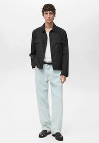 Zwarte, knoopjack met twee borstzakken, gedragen over een wit shirt. Lichtblauwe jeans en zwarte sandalen maken de outfit compleet.