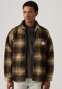 Chaqueta de forro polar a cuadros en tonos marrón y beige, con un forro de mezclilla gris, que cuenta con un bolsillo en el pecho y una pequeña etiqueta de logo. Textura suave, corte relajado.