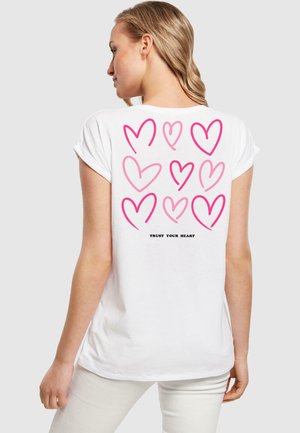 Weißes T-Shirt mit hochgekrempelten Ärmeln, das auf der Rückseite ein Design aus rosa Herzen und den Text "VERTRAU DEINEM HERZEN" in Schwarz zeigt. Hergestellt aus weicher Baumwolle.
