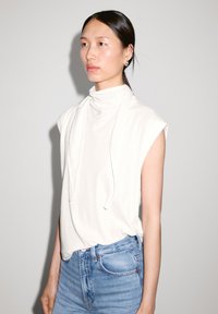 White sleeveless top with a high neck and draped detail, paired with light blue jeans. Smooth fabric, relaxed fit, minimalist design.