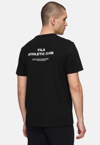 Musta puuvillainen T-paita, jossa on suuri valkoinen teksti "FILA ATHLETIC CLUB" selässä. Rentoa mitoitusta, lyhyet hihat ja pyöreä pääntie.
