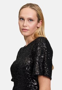 Schwarzes Paillettenoberteil mit kurzen Ärmeln. Verfügt über einen runden Ausschnitt und eine glatte, glänzende Textur. Figurnaher Schnitt mit funkelnenden Akzenten.