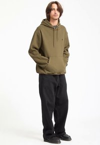 Olivovo zelená mikina s kapucňou a predným vreckom, doplnená o čierne voľné nohavice a čierne topánky.