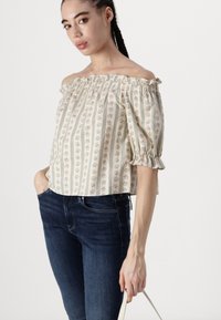 Off-shoulder blus i krämfärg med blå blommönster och vertikala ränder, med puffärmar och elastisk halsringning. Bärs med mörka jeans.