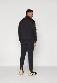 Black sweatshirt with high collar, back detailing, and graphic print on left sleeve. Paired with dark pants and beige athletic shoes.