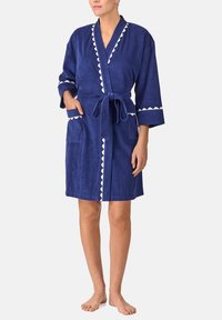 Donna che indossa un accappatoio blu navy lungo fino al ginocchio, con bordo bianco a scallop, tasche frontali e cintura annodata, in piedi scalza su uno sfondo bianco.