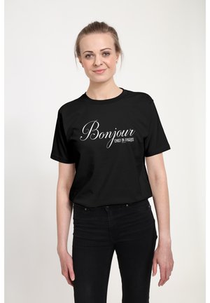Vrouw met licht haar in een staart, gekleed in een zwart T-shirt met witte tekst "Bonjour Emily in Paris", gecombineerd met een zwarte broek.