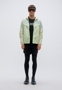 Ελαφρύ, διαφανές πράσινο μπουφάν με γκρι λεπτομέρειες, σε συνδυασμό με μαύρο μπλουζάκι, μαύρες βερμούδες και μαύρα κολάν. Φοράει ανθεκτικά αθλητικά παπούτσια.