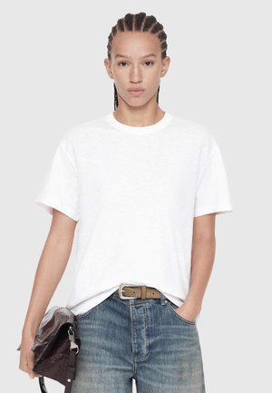 Person with braided hair wearing a white t-shirt and blue jeans, holding a folded dark leather bag in one hand, standing against a plain background.