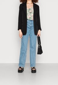Blazer noir sur un top floral vert, jeans taille haute bleus et sandales à plateforme noires. Elle tient un petit sac à main noir.