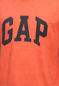 Tissu orange avec de grandes lettres majuscules noires formant GAP sur le devant d’un vêtement.