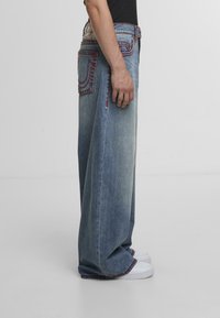 Osoba nosząca szerokie, jasnoniebieskie dżinsy z czerwonymi detalami oraz białe trampki, stojąca na tle jasnego, gładkiego tła.