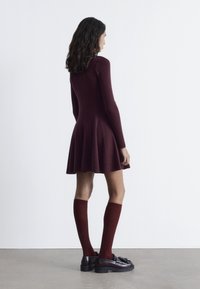 Burgunderfarbendes Rippkleid mit langen Ärmeln und schwingendem Rock. Das Model trägt passende knielange Socken und schwarze Loafer mit dekorativer Hardware.