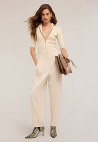 Kobieta z długimi falowanymi włosami nosi beżowy top na zamek, jasnobeżowe spodnie z wysokim stanem, botki z wężowym wzorem, duże kolczyki w kształcie kół oraz trzyma beżową kopertówkę.