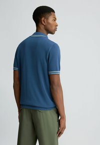 Blaues Strick-Polohemd mit kurzen Ärmeln, das helle blaue Streifen an den Bündchen und dem Kragen aufweist. Kombiniert mit grünen Hosen.