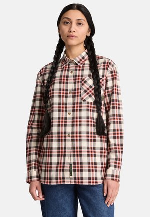 Ung person med langt flet hår iført en beige, rød og sort ternet skjorte med knapper og blå jeans mod en ensfarvet baggrund.
