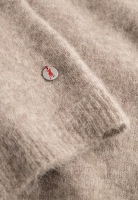 Beige wollen trui met een zachte textuur, voorzien van een ronde metalen tag met een rode dieren silhouet. Ribgebreide manchetten voegen detail toe.