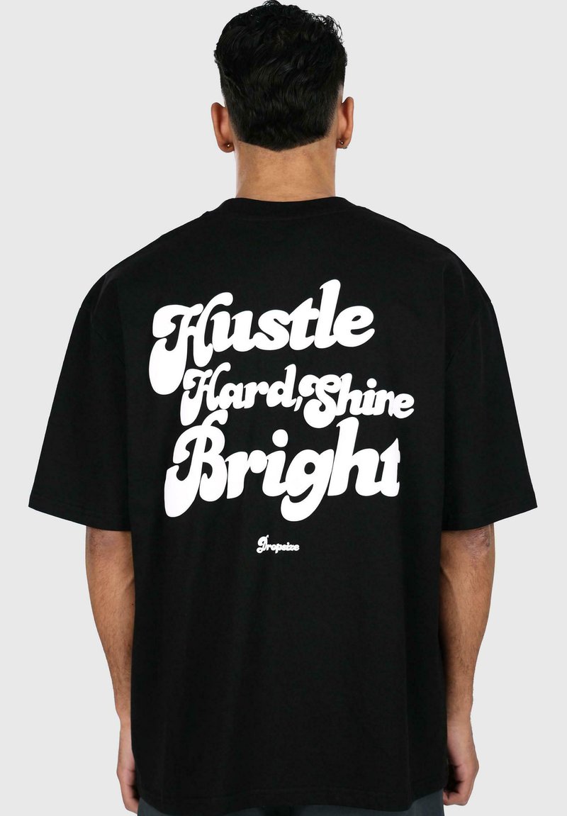 Sort bomulds t-shirt med løs pasform, prydet med store hvide bogstaver på ryggen, hvor der står "Hustle Hard, Shine Bright." Enkel design.