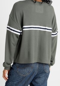 Grön ribbad tröja med lös passform, försedd med horisontella marinblå och vita ränder över ryggen. Matchad med högmidjade denimjeans.