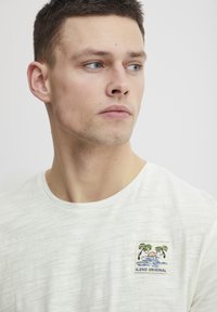 Heled hall t-särk tekstuuriga viimistlusega, millel on tikitud plekk palmi puudest ja veest, millel on silt "BLEND ORIGINAL."