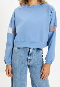 Světle modrý zkrácený svetr s dlouhými rukávy, který má průhledné síťované panely na rukávech a elastické manžety. Jednoduchý design, měkká textura.