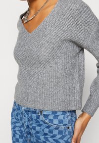 Grauer Strickpullover mit V-Ausschnitt, der über ein geripptes Muster und eine taillierte Silhouette verfügt, kombiniert mit blauen karierten Jeans.
