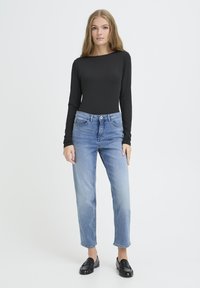 Žena stojaca, oblečená v čiernom tričku s dlhým rukávom, svetlomodrých džínsoch a čiernych mokasínach, na jednoduchom bielom pozadí.