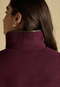 Sieviete ar gariem brūniem matiem, kura valkā zelta auskarus gredzenā un bordo flīsa jaku ar uzrakstu "JOULES" augstajā apkaklē.