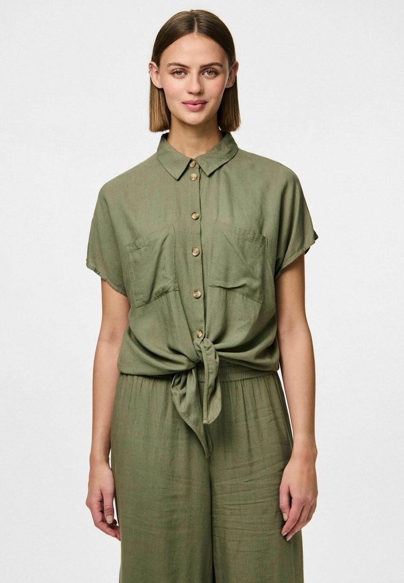 Chemise vert olive à manches courtes avec un devant boutonné, deux poches poitrine et un ourlet noué. Fabriquée en tissu léger et texturé.