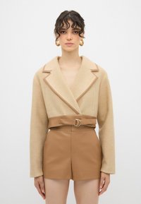 Beige tekstureret jakke med bred krave og brun bæltet, parret med brune shorts i kunstlæder. Designet har vinkelformede lommer og minimal syning.