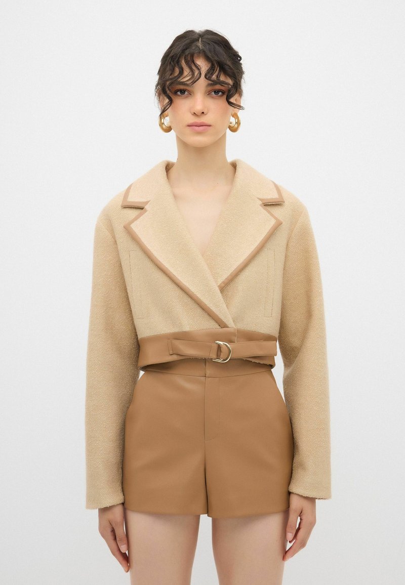 Beige tekstureret jakke med bred krave og brun bæltet, parret med brune shorts i kunstlæder. Designet har vinkelformede lommer og minimal syning.