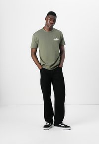 Olive green cotton t-shirt with a round neckline and small logo on the chest, paired with black straight-leg jeans and black sneakers.