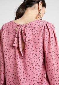 Roze blouse met een textuurstof met kleine zwarte bloemenpatronen, pofmouwen en een striksluiting aan de achterkant van de hals.