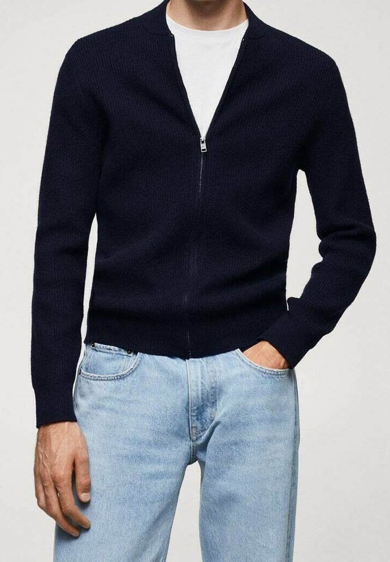 Homme portant un pull zippé bleu marine sur un t-shirt blanc avec un jean bleu clair, une main dans la poche, debout devant un fond uni.