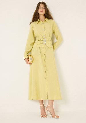 Femme portant une longue robe jaune boutonnée avec une ceinture à double boucle, tenant une pochette ronde dorée, et des sandales blanches à talons hauts contre un mur blanc.